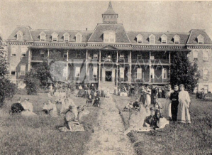 Sepia toned photograph of a building, and many women posed on the lawn in front of it