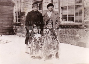 A sepia toned photograph of two adult women and two children posed for a picture outside. They are beside a stone house, there is snow on the ground, and they are all wearing winter clothes.