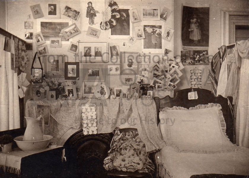Sepia photo of a room with a bed and several of photos on the wall. There is a washstand to the left