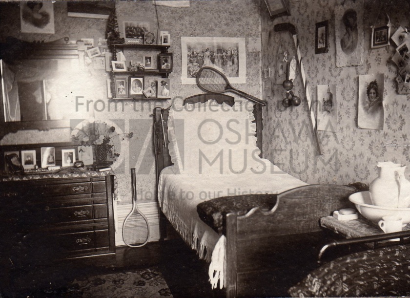 Sepia photo of a room with a bed and dresser. There are several photos around the room, with two tennis rackets and a hockey stick