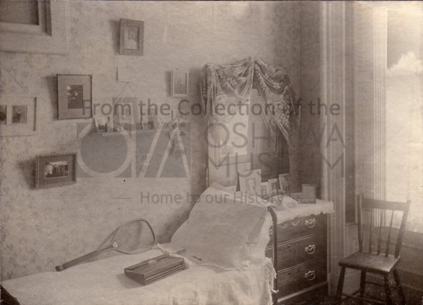 Sepia photo of a room with a bed and dresser. There are a few photos on the room, a chair, and a tennis racket on the bed