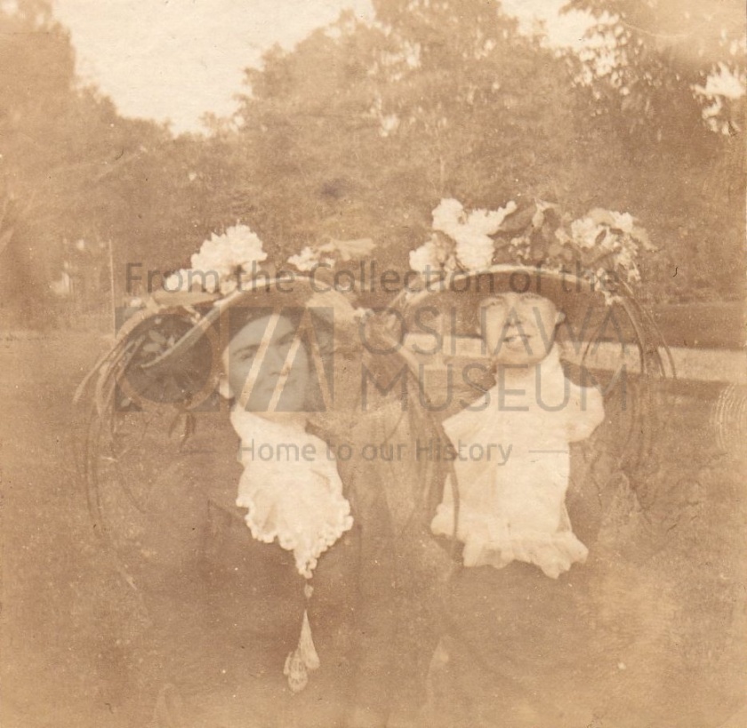 Sepia photo of two Caucasian women, seated, wearing large floral hats.