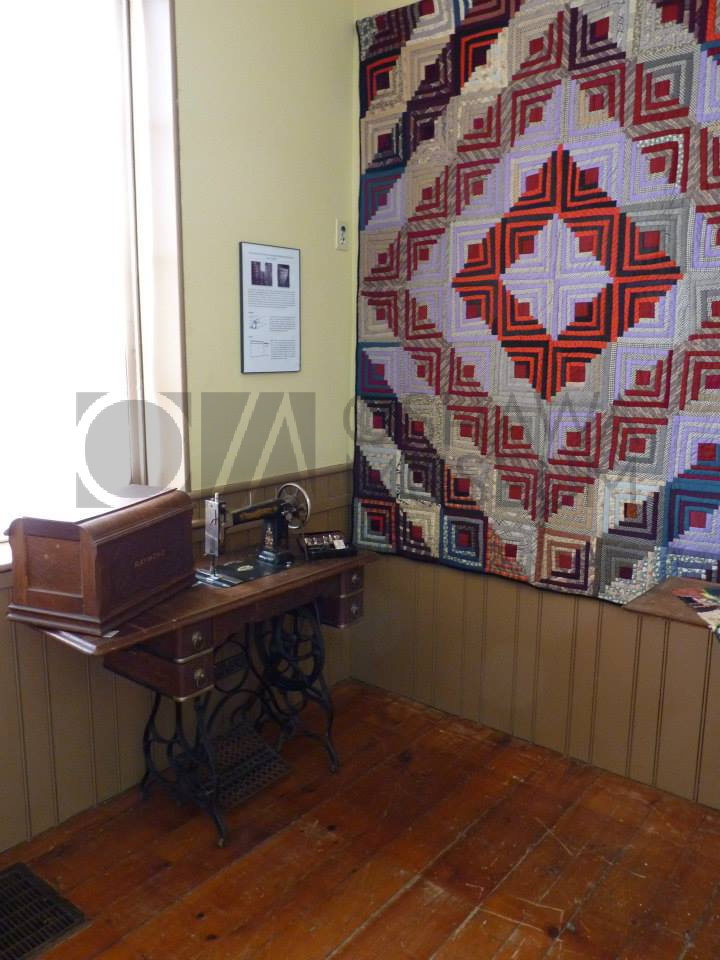 A room with a colourful quilt hanging from the wall and a wooden and metal sewing machine in front of it