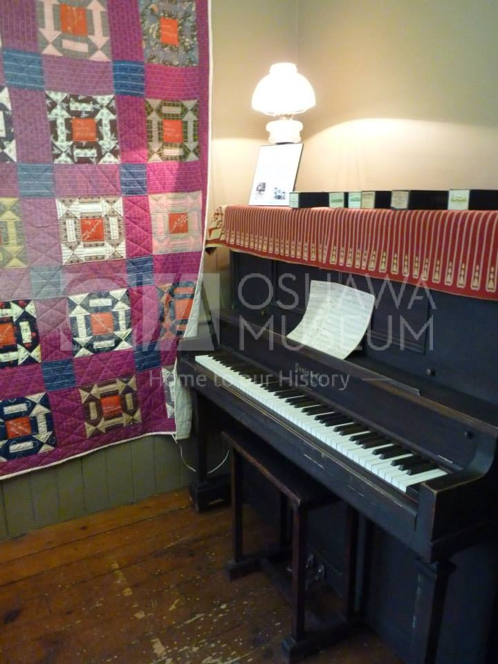 A room with a colourful quilt hanging on the wall. A large, wooden piano is beside the quilt.