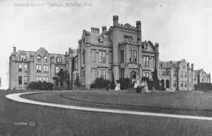 Black and white photo of a large, palatial building. There is a group of people on the lawn in front of the building.